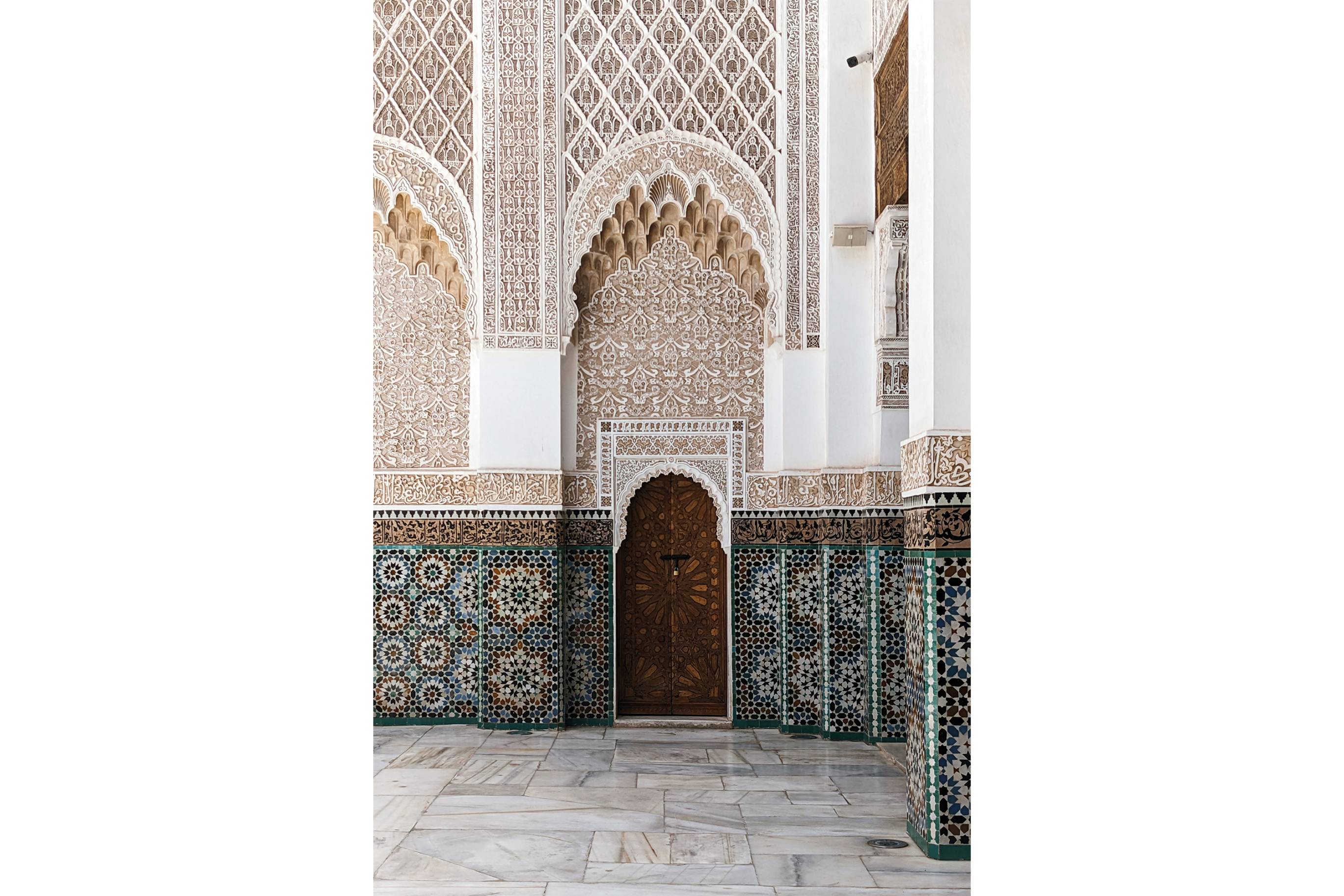 Ornate wooden door in the middle of a Zellij and plaster carving covered wall of the courtyard of the Madrassa Ben Youssef in Marrakech, Morocco