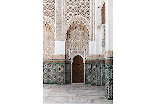 Ornate wooden door in the middle of a Zellij and plaster carving covered wall of the courtyard of the Madrassa Ben Youssef in Marrakech, Morocco