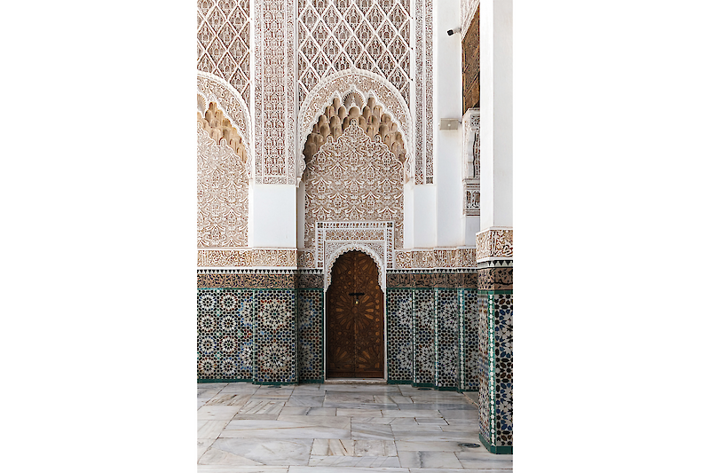 Ornate wooden door in the middle of a Zellij and plaster carving covered wall of the courtyard of the Madrassa Ben Youssef in Marrakech, Morocco